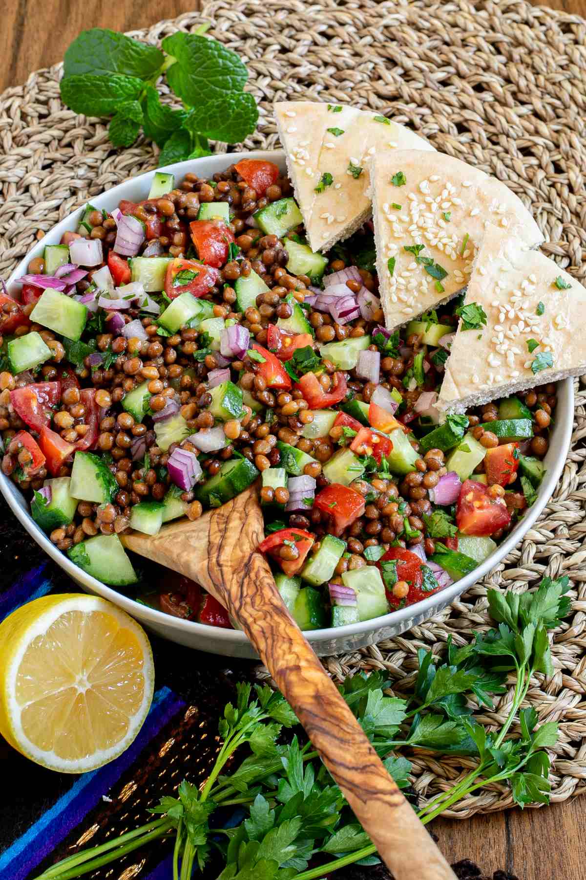 Large white bowl with a chopped mixed salad of brown lentils, cucumber, tomatoes, and red onion. 3 pita triangles are placed on the side. A wooden spoon is placed inside.