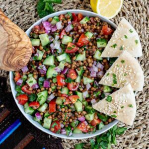 Large white bowl with a chopped mixed salad of brown lentils, cucumber, tomatoes, and red onion. 3 pita triangles are placed on the side.