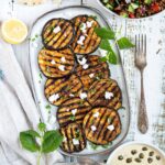 Large grilled slices of eggplant sprinkled with fresh herbs and crumbled cheese served on a light blue plate.