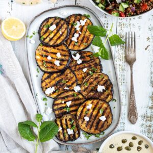 Large grilled slices of eggplant sprinkled with fresh herbs and crumbled cheese served on a light blue plate.