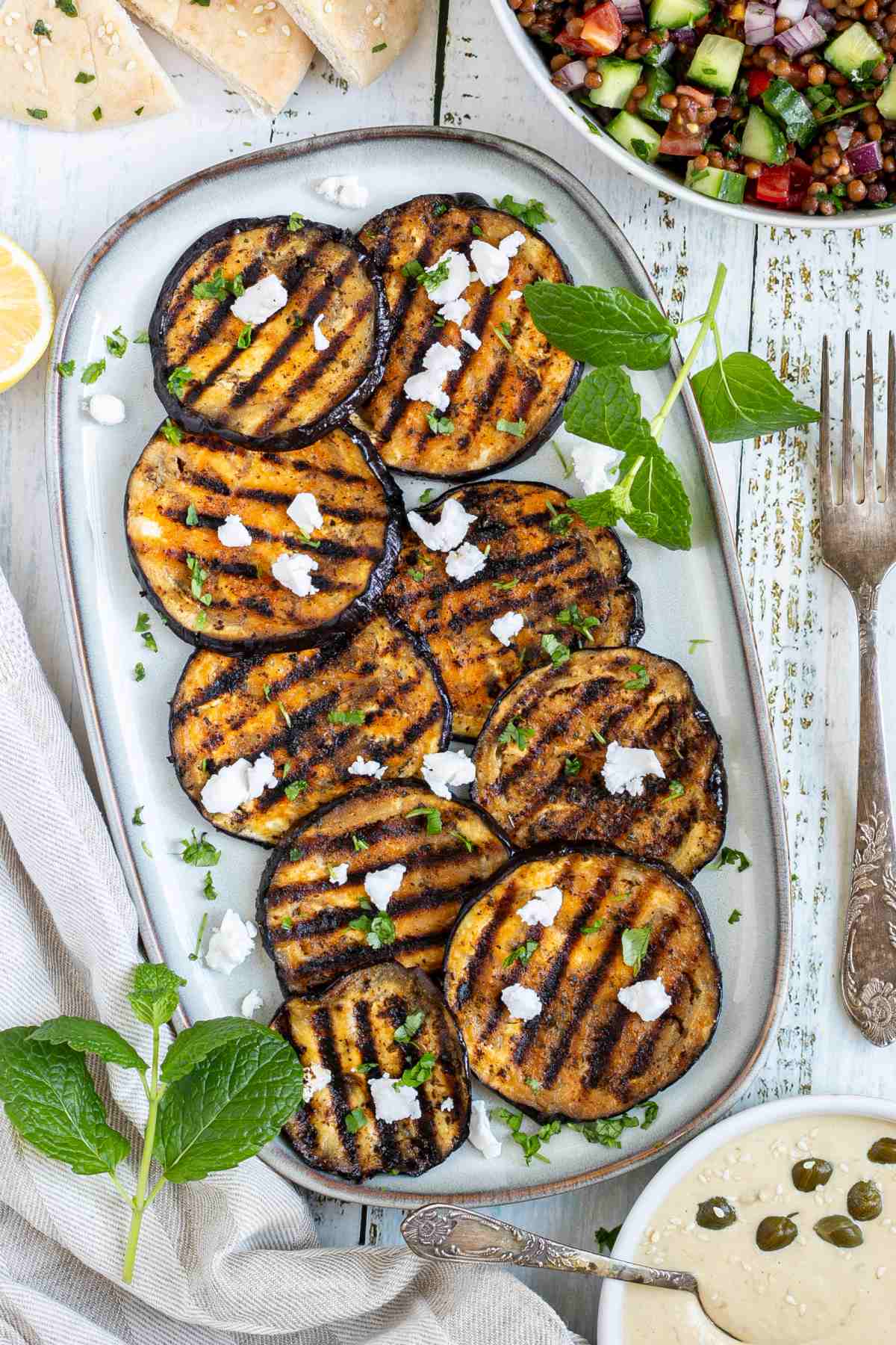 Large grilled slices of eggplant sprinkled with fresh herbs and crumbled cheese served on a light blue plate.