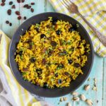 Yellow rice with raisins and pistachios and green herbs in a black bowl. More dry ingredients are scattered around it.