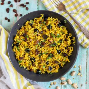 Yellow rice with raisins and pistachios and green herbs in a black bowl. More dry ingredients are scattered around it.