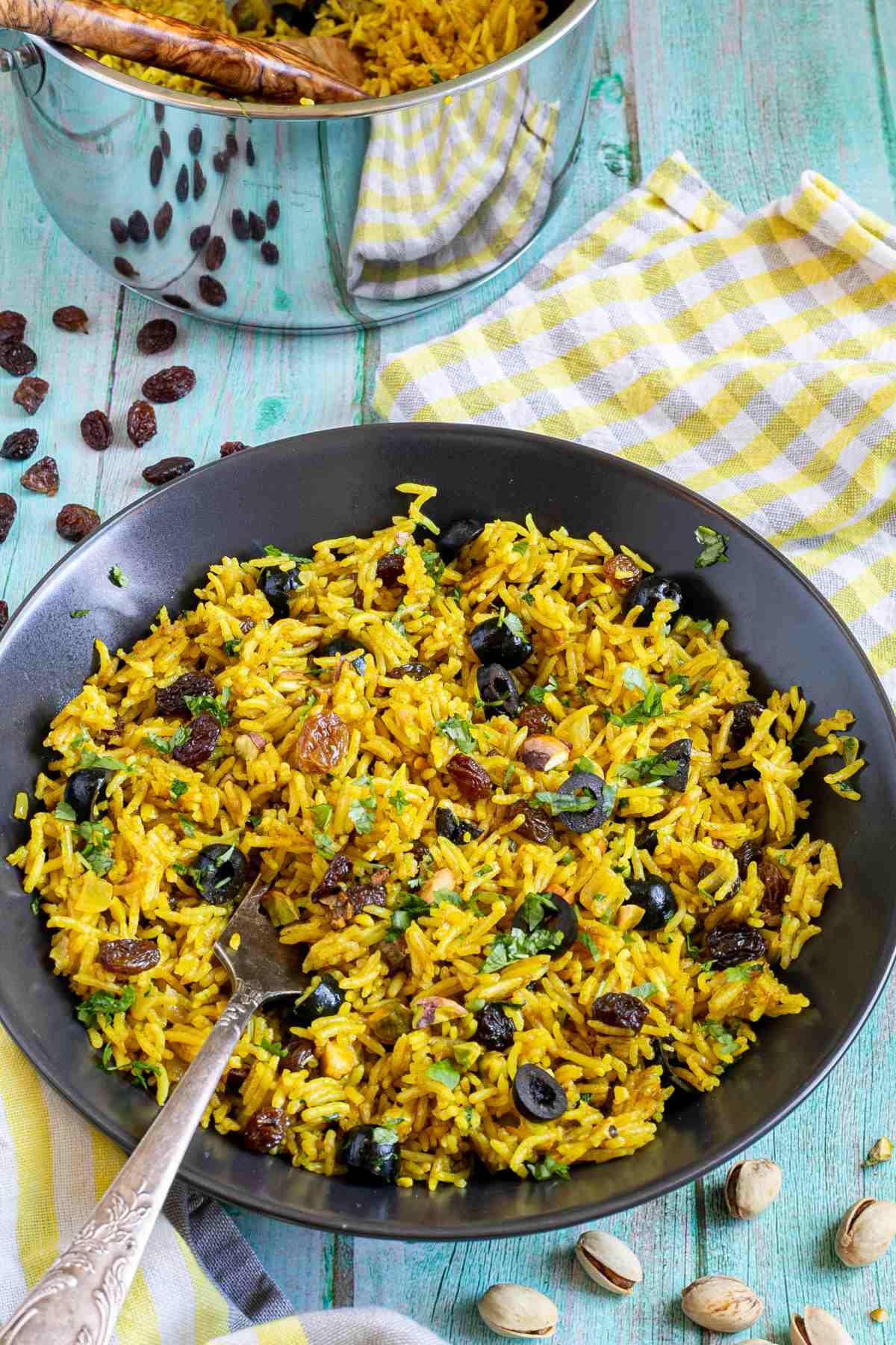 Yellow rice with raisins and pistachios and green herbs in a black bowl. More dry ingredients are scattered around it.