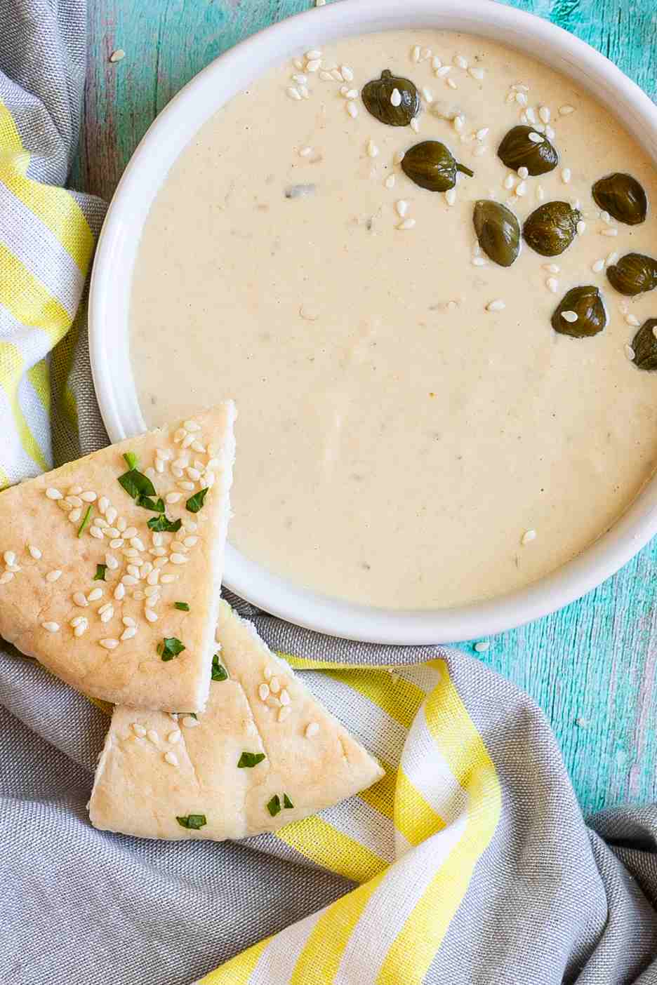 Small white bowl with a light brown thick sauce topped with sesame seeds and capers. Two pita triangles are placed on the side.