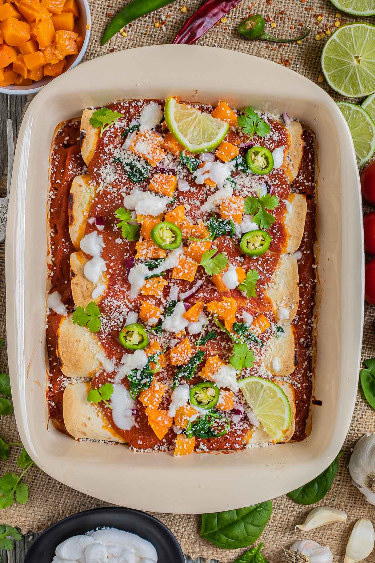 A casserole pan with rolled tortillas covered in red sauce and topped with diced roasted butternut squash, chili pepper slices, cilantro leaves, lime slices, sour cream and grated cheese.