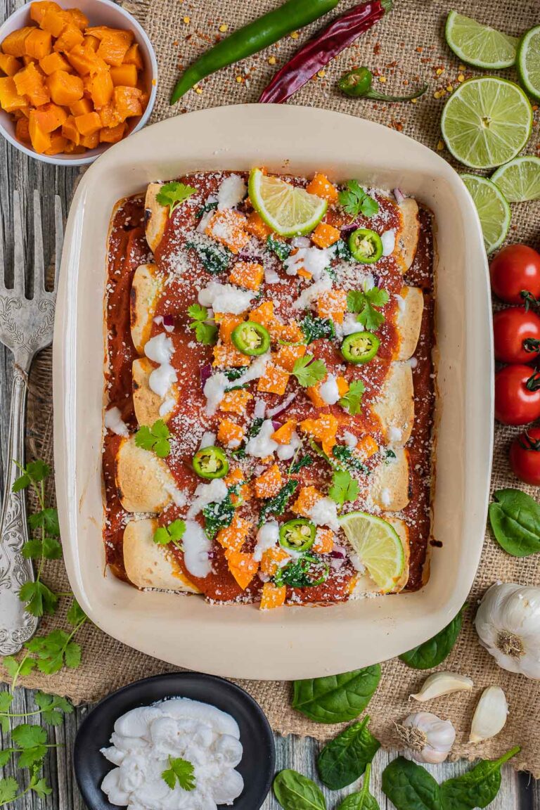 A casserole pan with rolled tortillas covered in red sauce and topped with diced roasted butternut squash, chili pepper slices, cilantro leaves, lime slices, sour cream and grated cheese. More ingredients in small bowls right next to it.