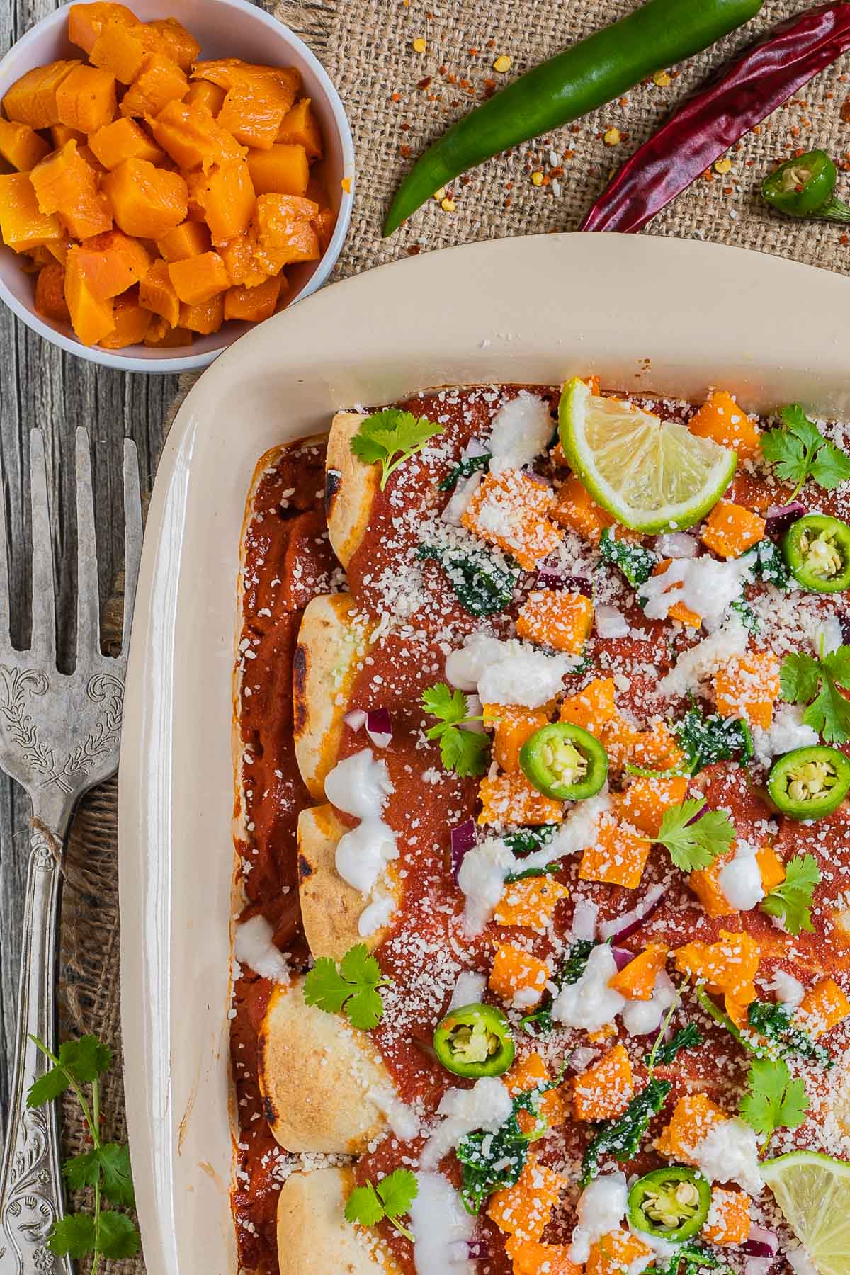 A casserole pan with rolled tortillas covered in red sauce and topped with diced roasted butternut squash, chili pepper slices, cilantro leaves, lime slices, sour cream and grated cheese. A small bowl of diced roasted squash is next to it.