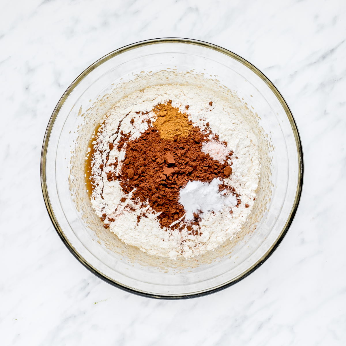 A glass bowl with brown liquid, flour, cinnamon, and cocoa powder.
