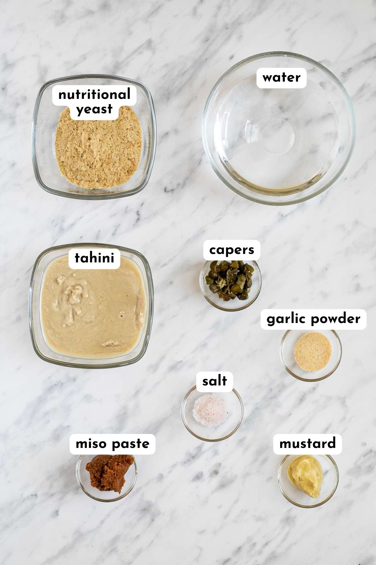 Ingredients of tahini caesar dressing in small glass bowls like water, tahini, yellow flakes, capers, salt, dark brown paste, mustard, and garlic powder.  