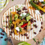 Two tortillas with charred grill lines topped with colorful bell pepper and onion strips as well as black beans, topped with sour cream and guacamole. Corn-based side dishes are in small bowls next to them.