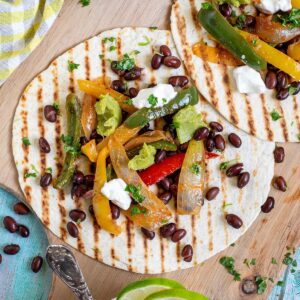 Two tortillas with charred grill lines topped with colorful bell pepper and onion strips as well as black beans, topped with sour cream and guacamole. Corn-based side dishes are in small bowls next to them.