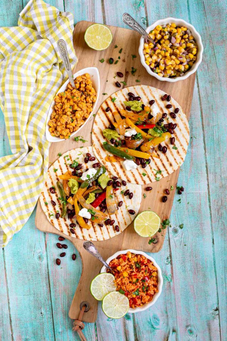 Two tortillas with charred grill lines topped with colorful bell pepper and onion strips as well as black beans, topped with sour cream and guacamole. Corn-based side dishes are in small bowls next to them.