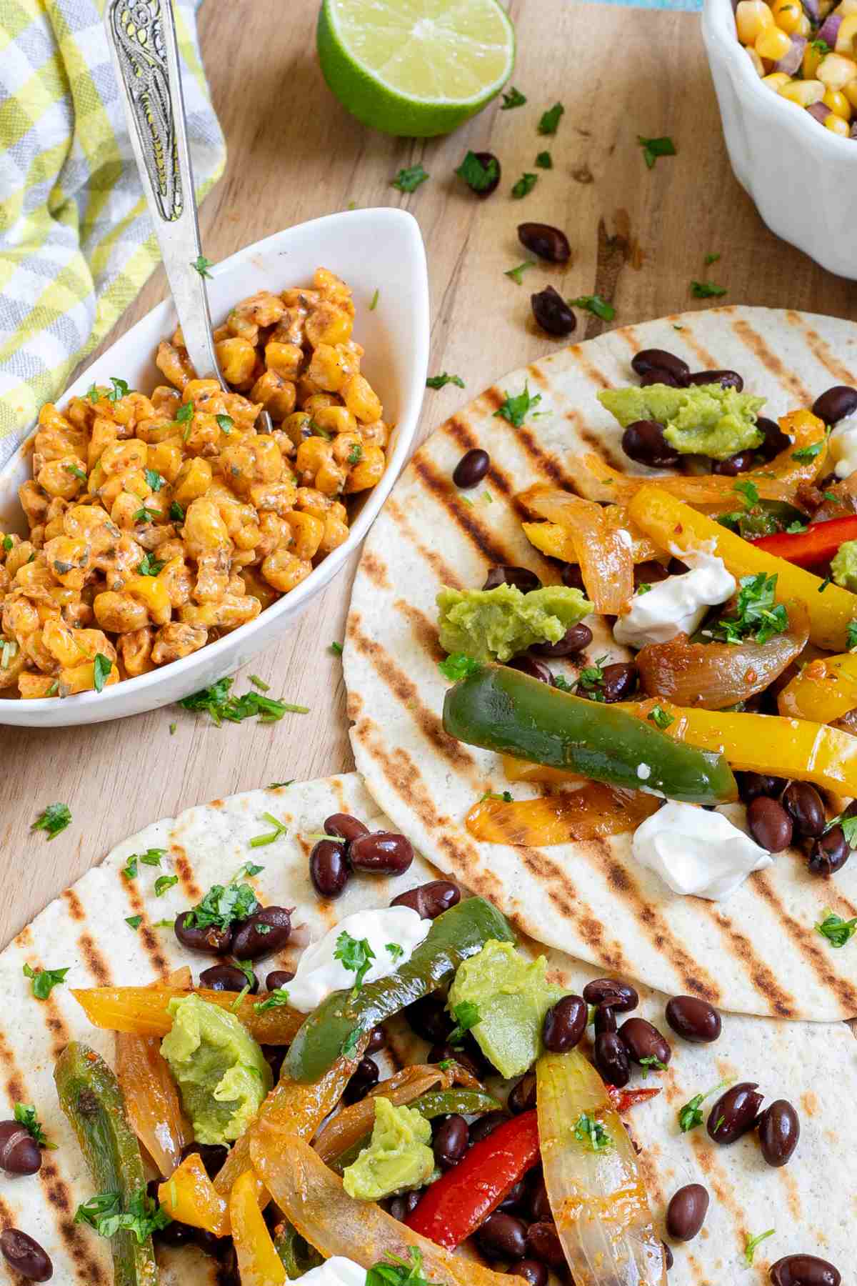 Two tortillas with charred grill lines topped with colorful bell pepper and onion strips as well as black beans, topped with sour cream and guacamole. Corn-based side dishes are in small bowls next to them.