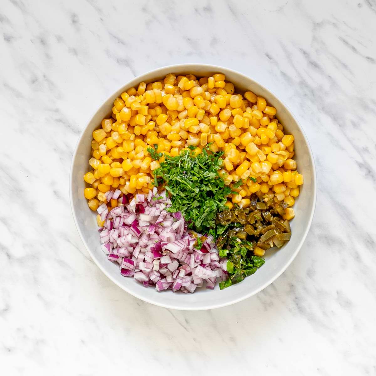 A large white bowl with sweet corn kernels, chopped red onion, chopped green herbs, and chopped green chilies.