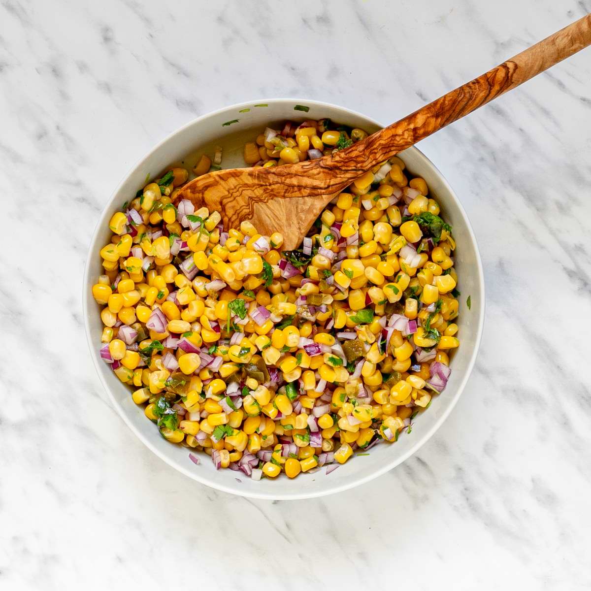 A large white bowl with a mix of sweet corn kernels, chopped red onion, chopped green herbs, and chopped green chilies.