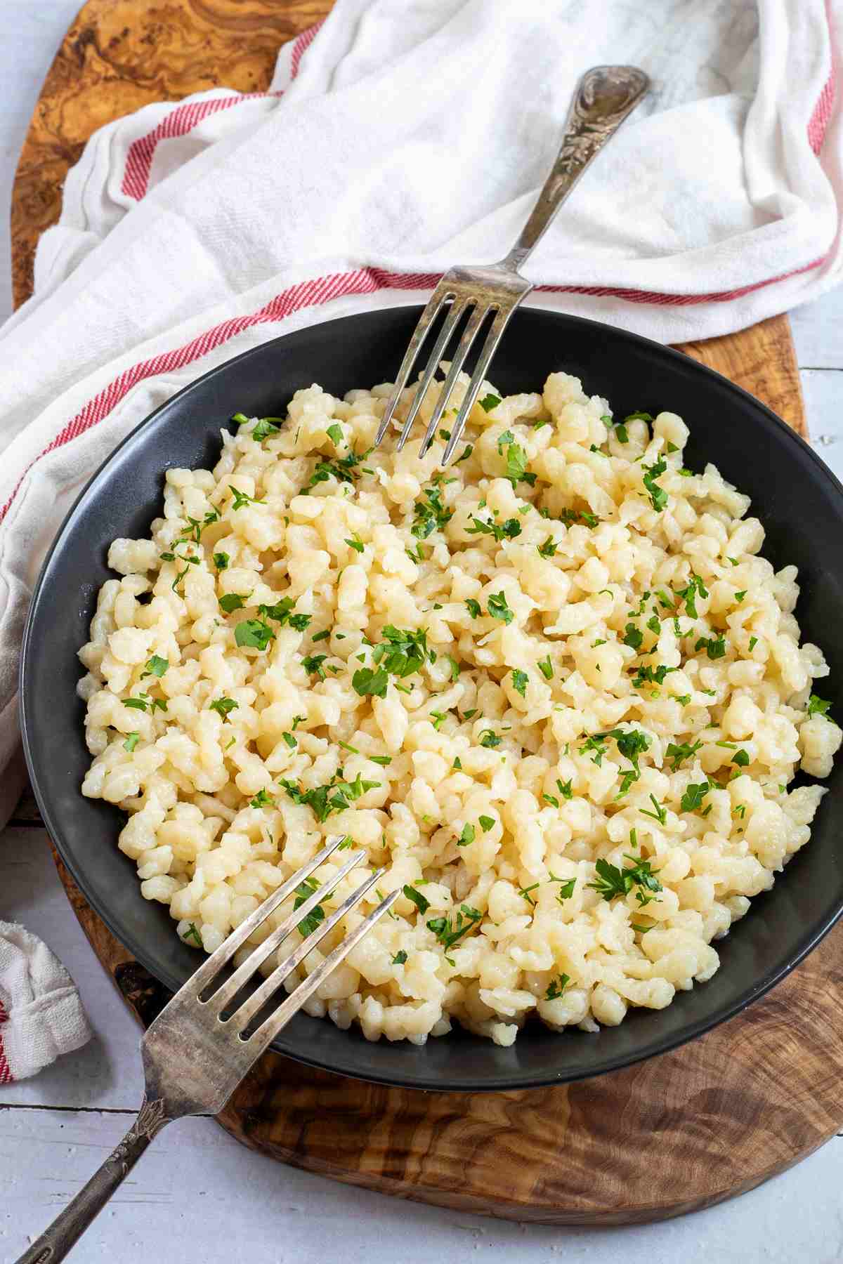 A bowl of Hungarian dumplings, also known as Nokedli or Csipetke, with chopped fresh parsley on a wooden cutting board with two forks.