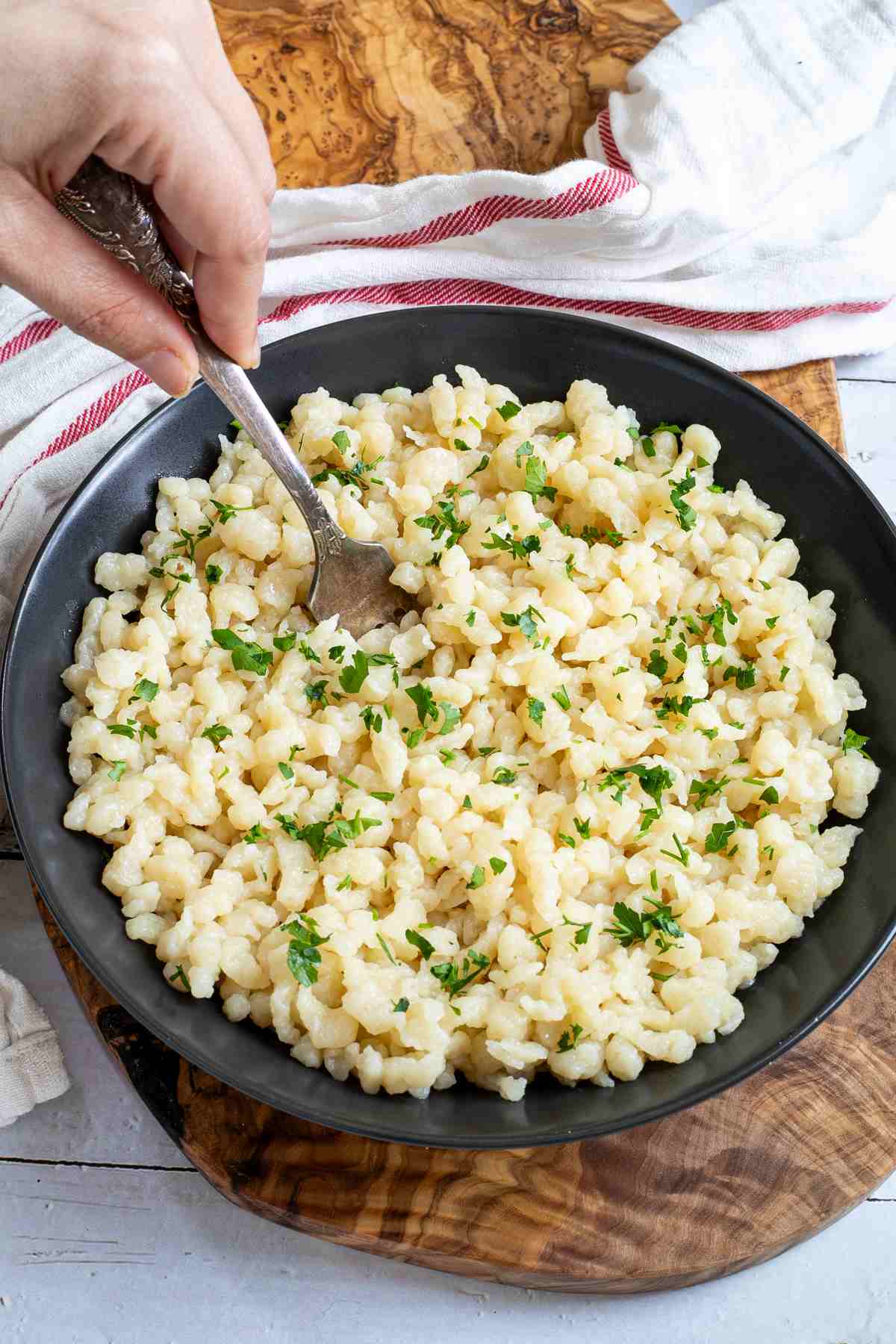 A person holding a fork in a bowl of Hungarian dumplings with chopped fresh parsley on a wooden cutting board.