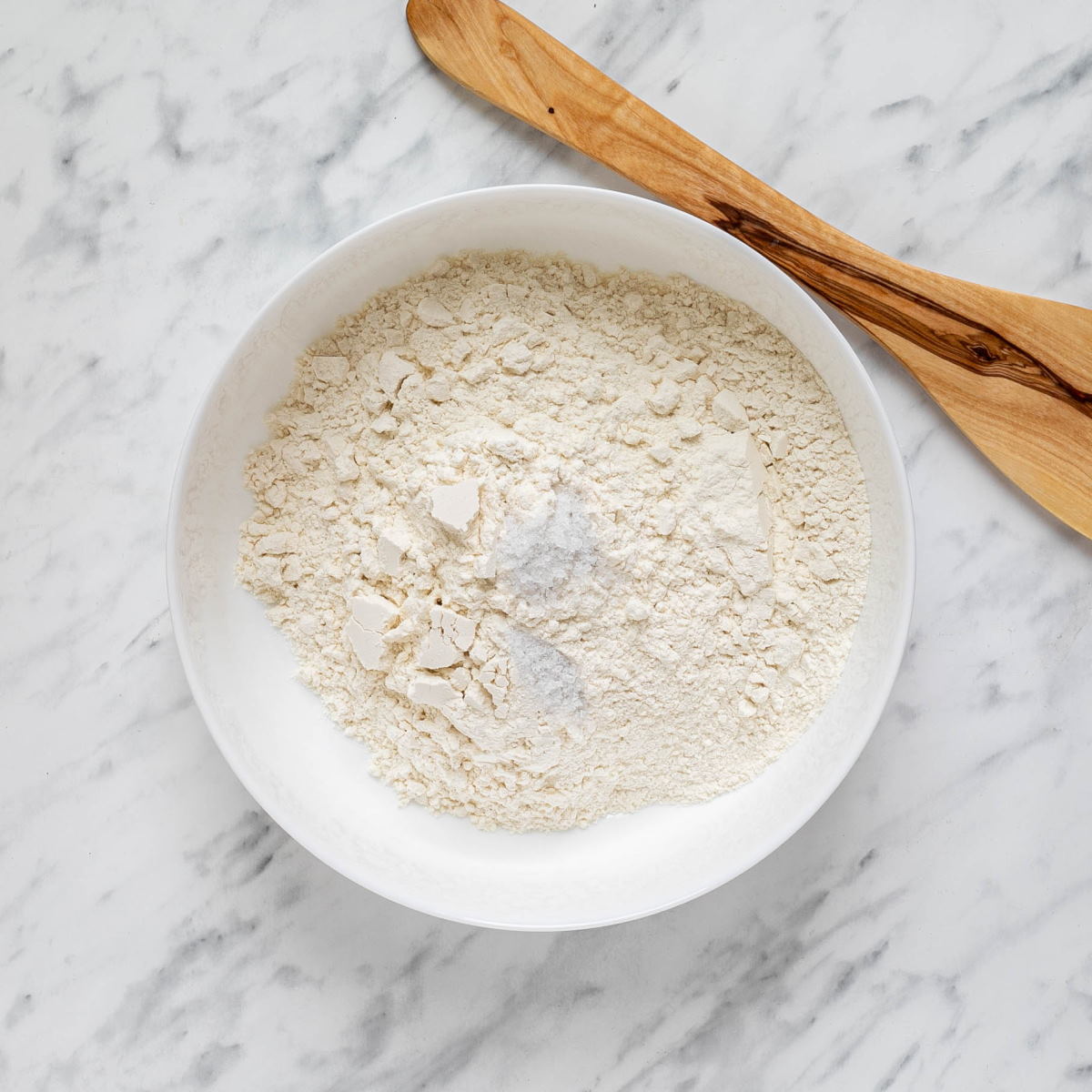 Dry ingredients to make Hungarian nokedli in a bowl on a white marble surface with a wooden spoon on the side.