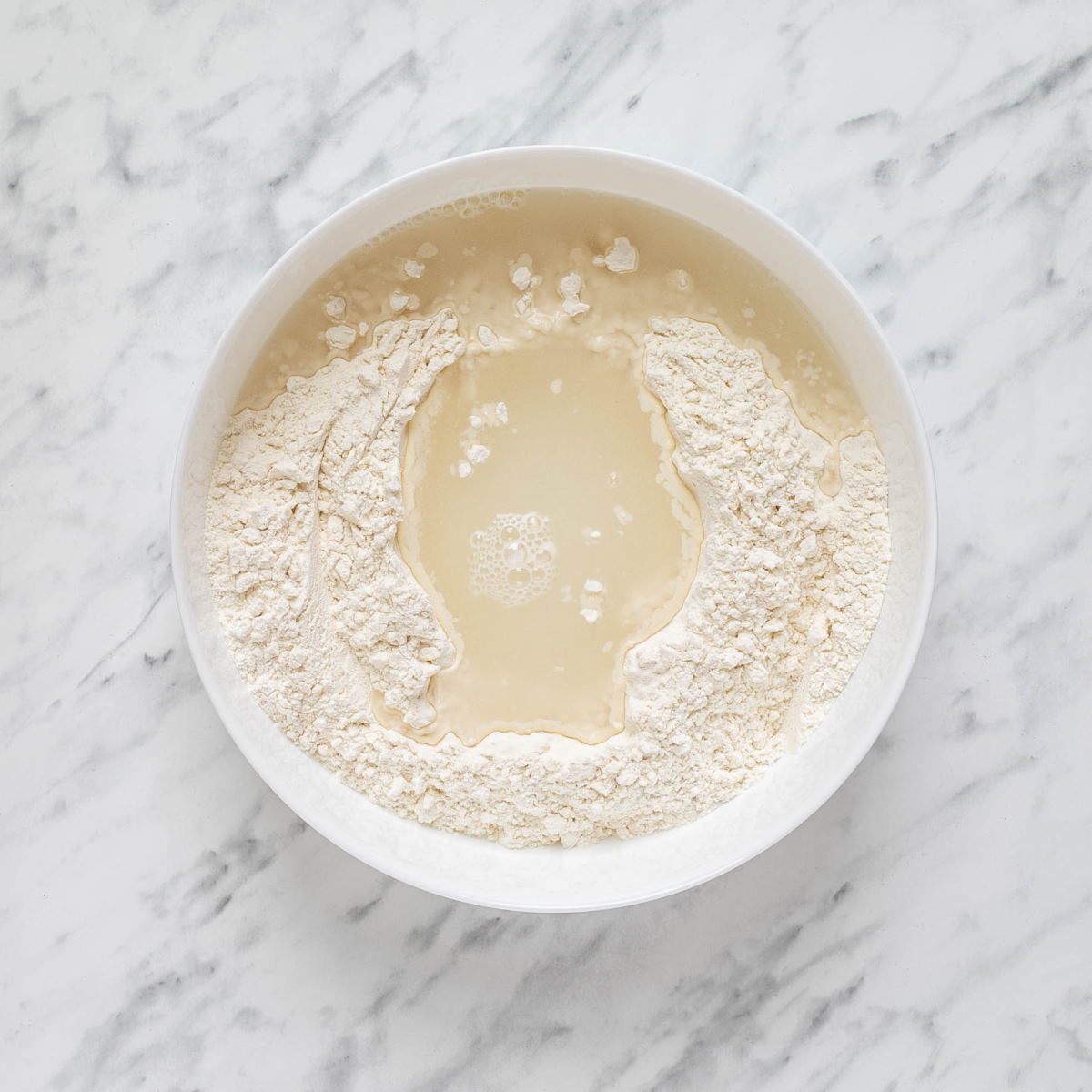 Dry ingredients with water to make Hungarian nokedli in a bowl on a white marble surface with a wooden spoon on the side.