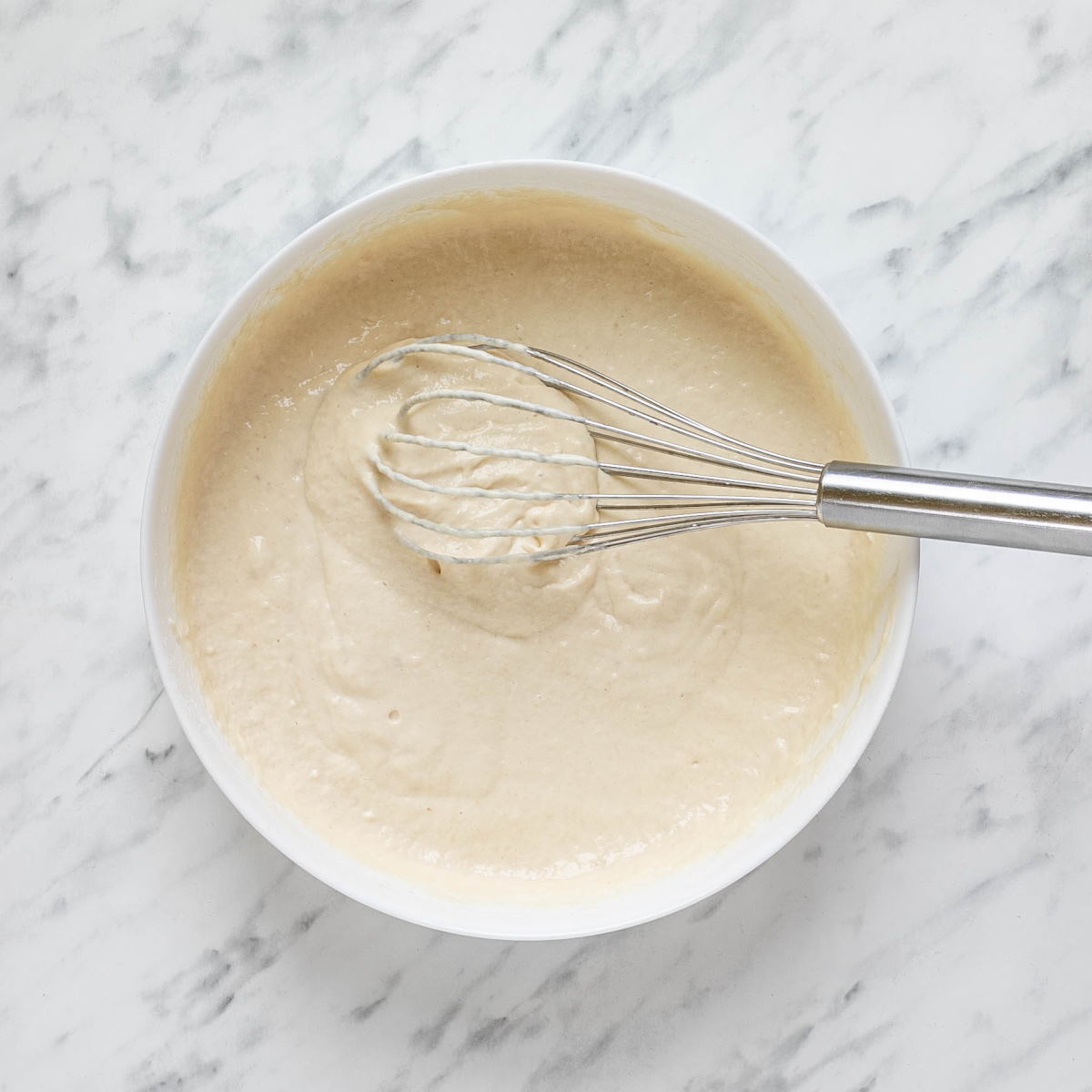 Hungarian nokedli batter in a bowl on a white marble surface with a wooden spoon on the side.