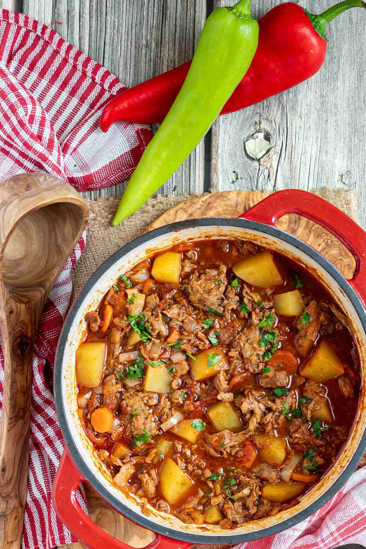 A pot of authentic Hungarian Goulash with meat and vegetables on a wooden table with two peppers on the side.