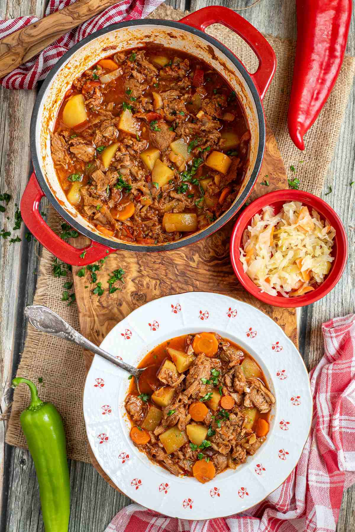 A pot of authentic Hungarian Goulash with meat and vegetables on a wooden table with a portion in a white plate on the side.
