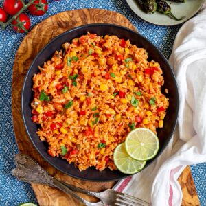 Orange rice with corn, tomato, and parsley in a black bowl. Lime slices are on the side.