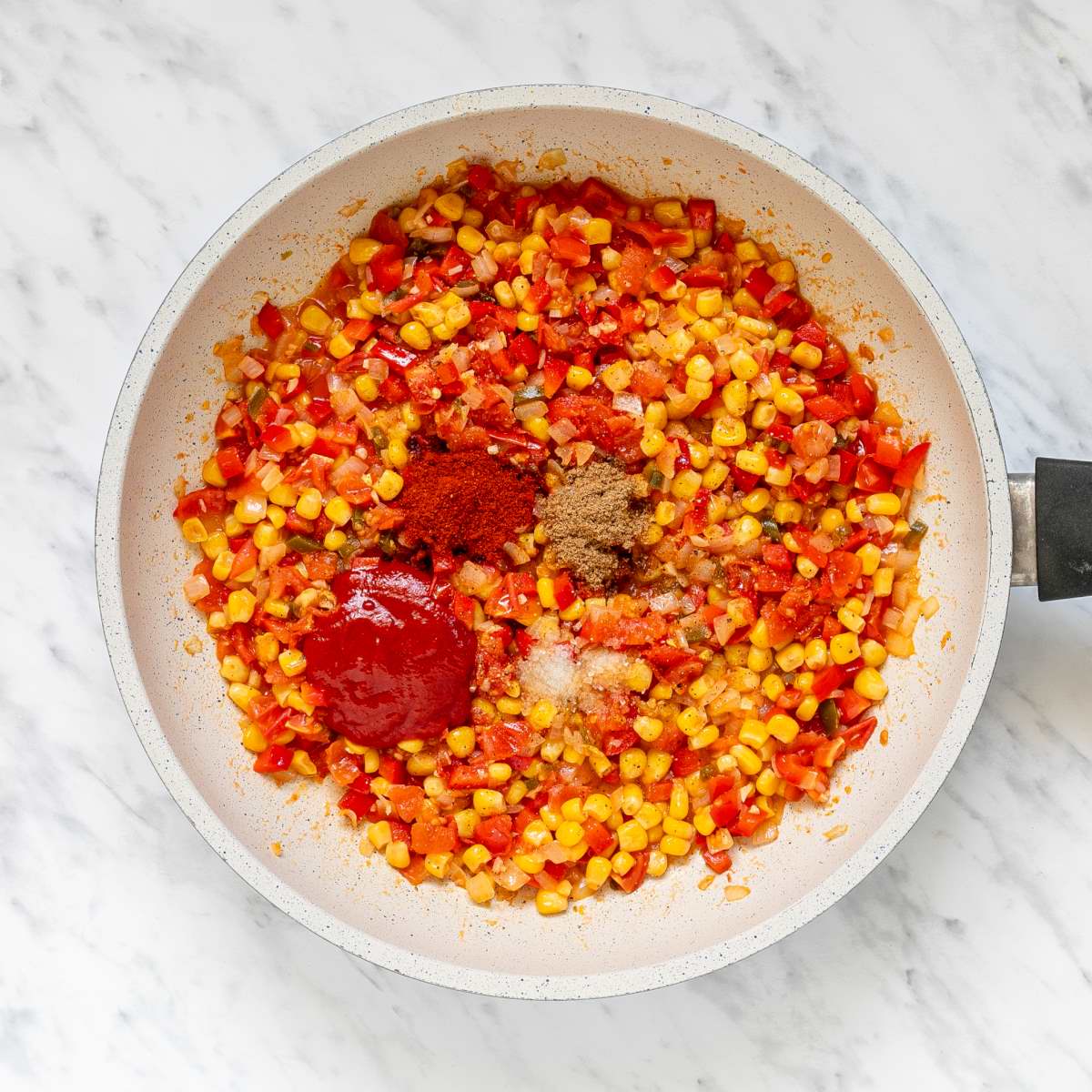 White frying pan with a mix of chopped tomatoes, bell peppers, corn and olives topped with small heaps of different spices.