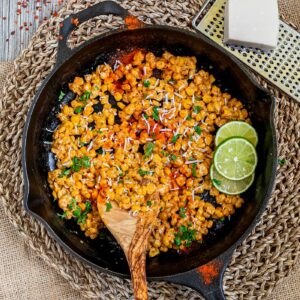 Cast iron skillet with charred yellow corn covered in spices and chopped herbs. A wooden spatula and two lime slices are placed inside.
