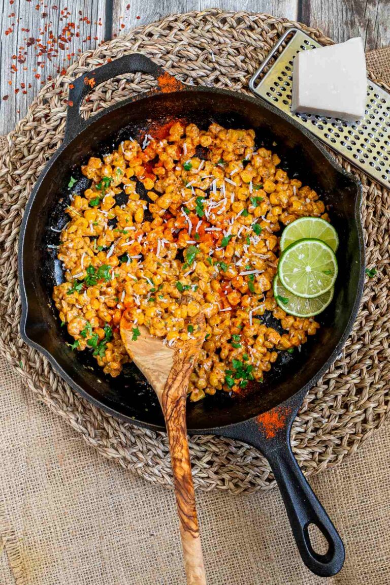 Cast iron skillet with charred yellow corn covered in spices and chopped herbs. A wooden spatula and two lime slices are placed inside.