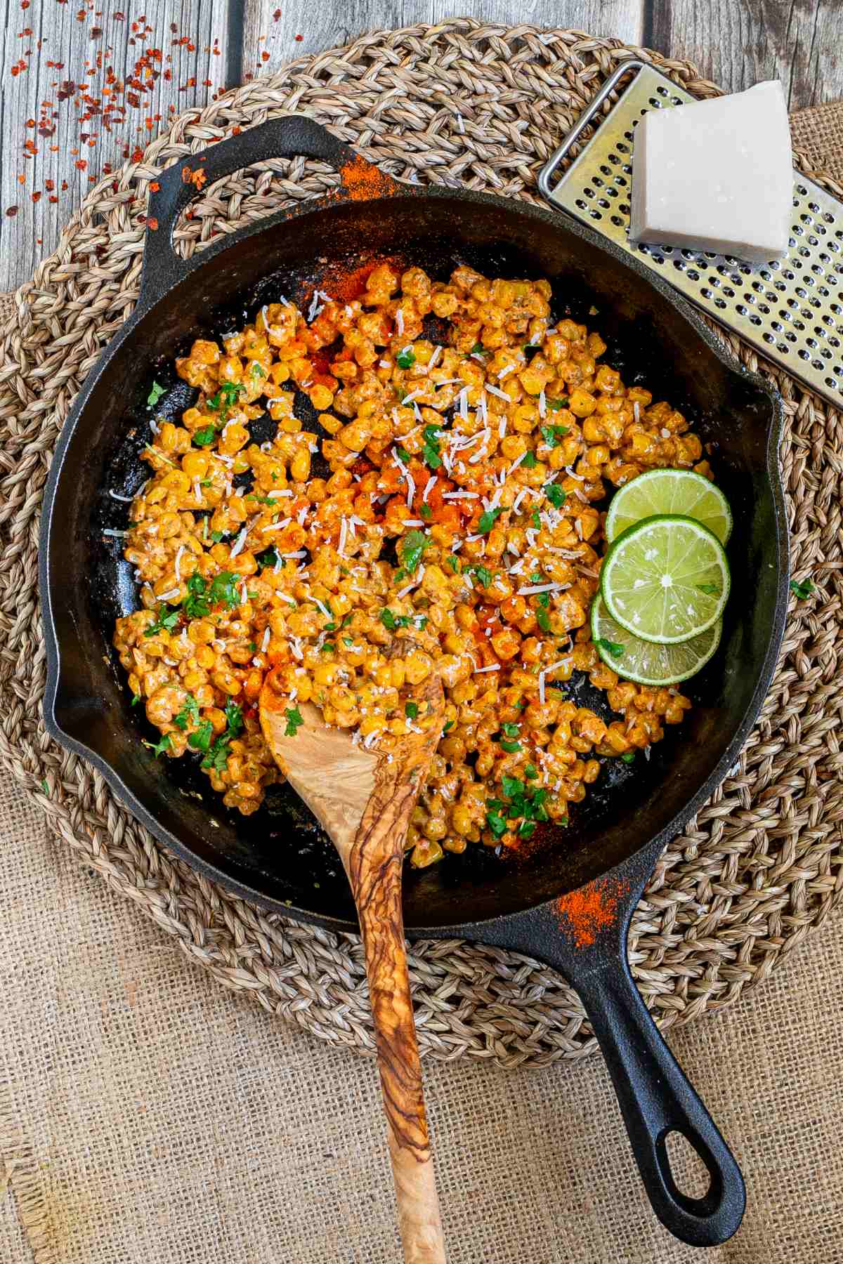 Cast iron skillet with charred yellow corn covered in spices and chopped herbs. A wooden spatula and two lime slices are placed inside. 