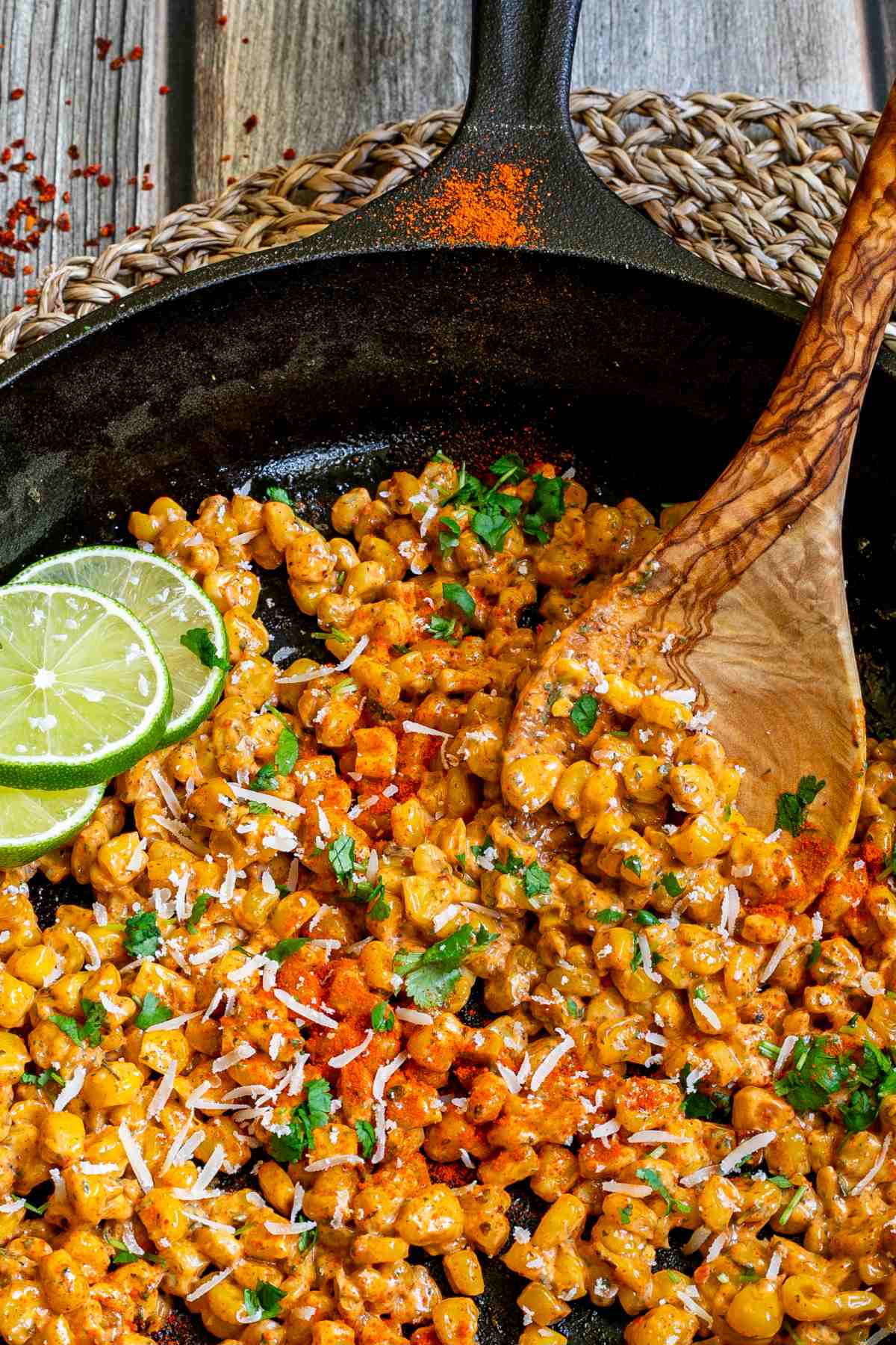 Cast iron skillet with charred yellow corn covered in spices and chopped herbs. A wooden spatula and two lime slices are placed inside.