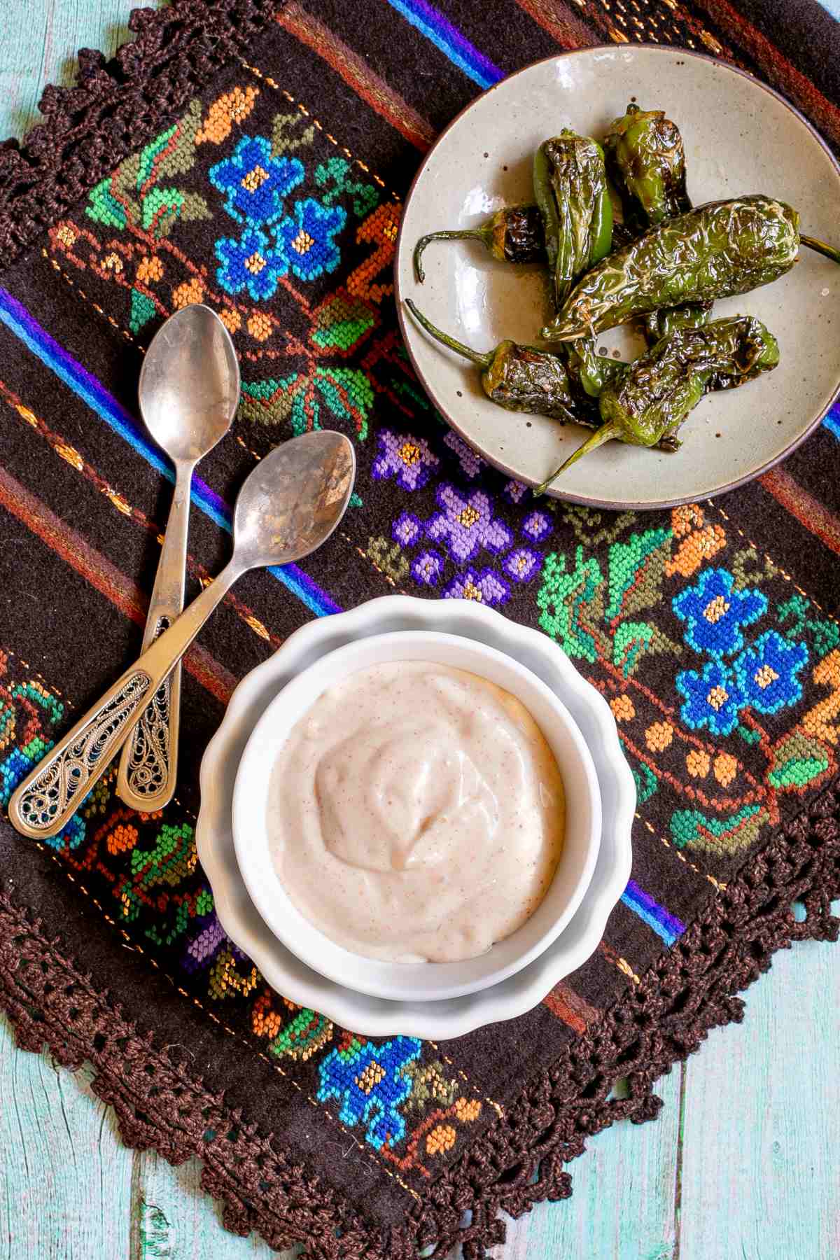 Light orange thick dip is in a small bowl with roasted green peppers on the side. Placed on a colorful brown, blue, green woven tablecloth.