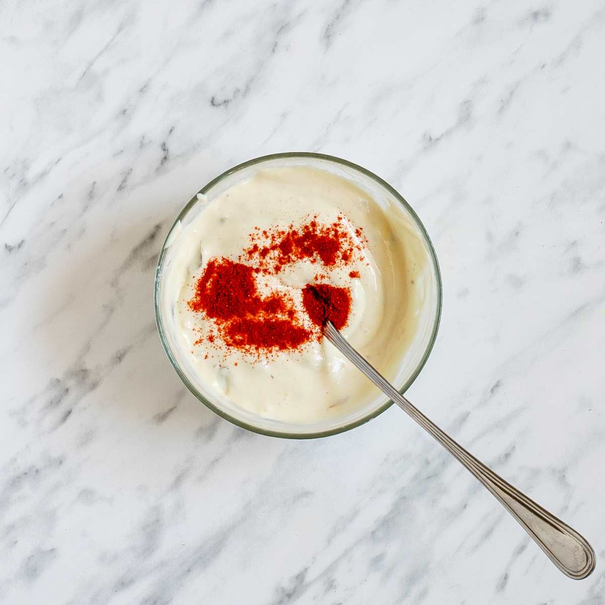 Small glass bowl with a spoon filled with thick white dip and a small heap of red paprika powder.