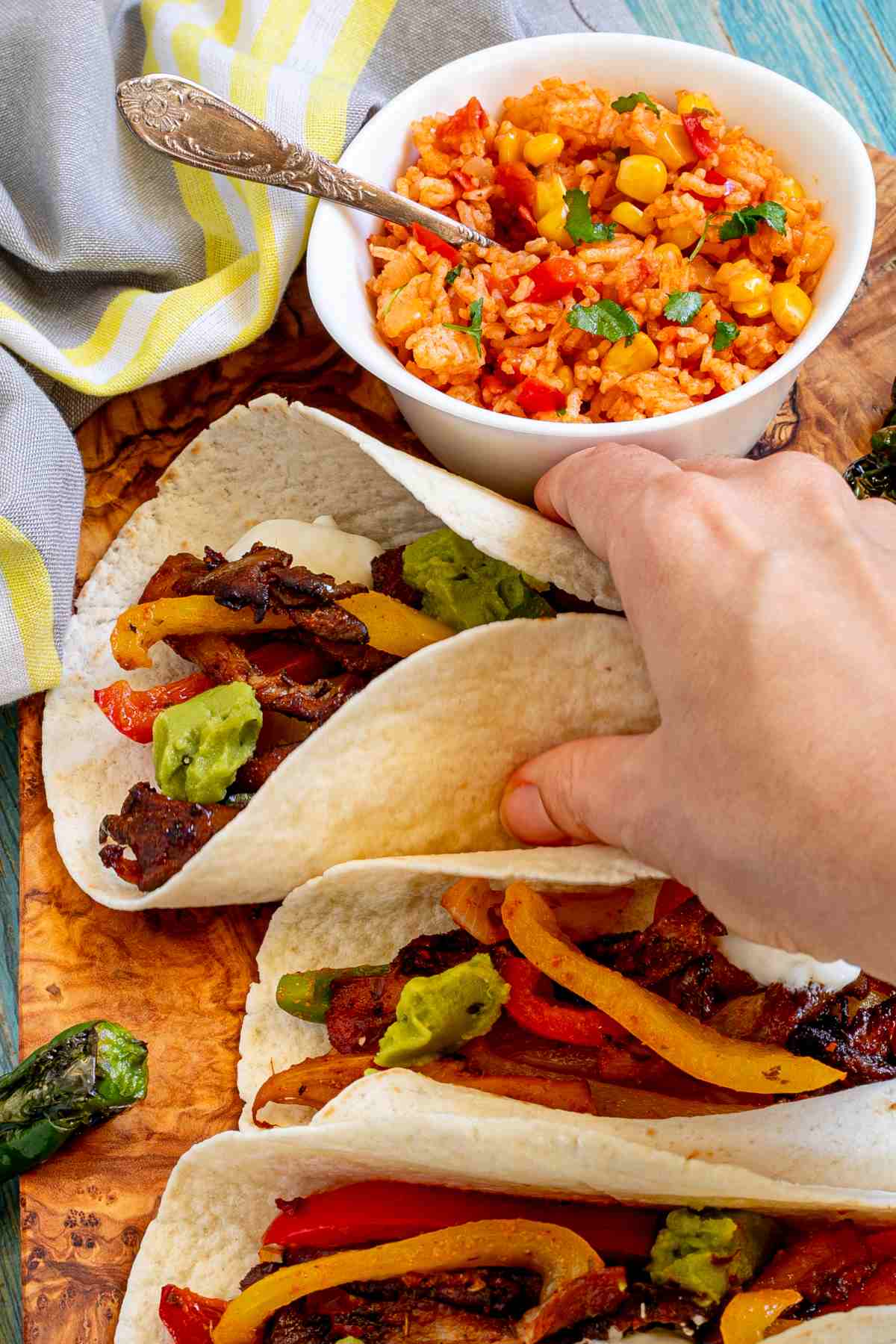 A hand is holding a small tortilla with colorful pepper slices and crispy brown mushroom shreds topped with white and green sauces. Surrounded by side dishes.