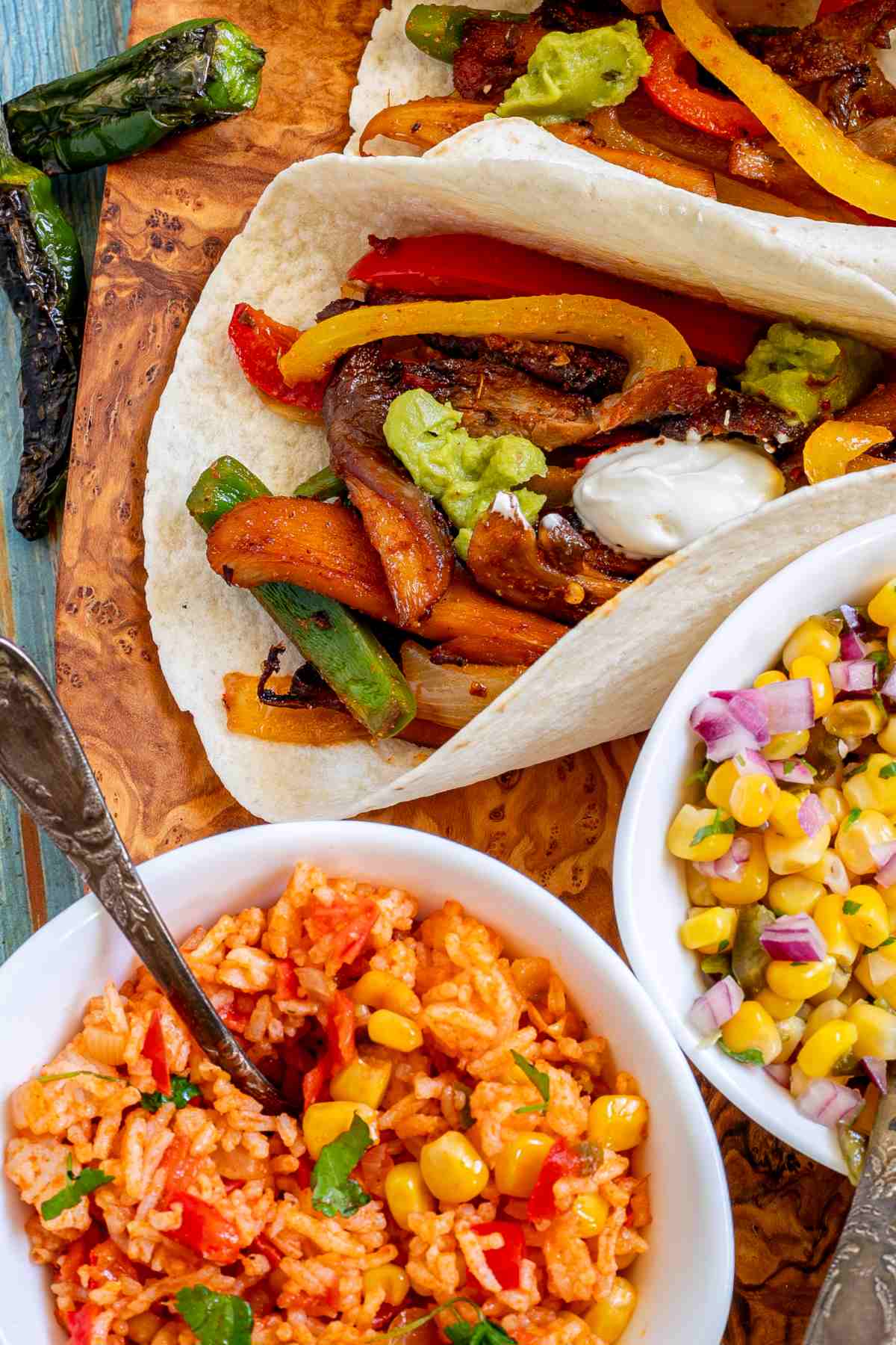 Small tortillas with colorful pepper slices and crispy brown mushroom shreds topped with white and green sauces on a wooden board. Surrounded by side dishes.