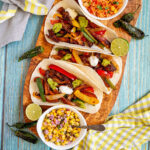 Small tortillas with colorful pepper slices and crispy brown mushroom shreds topped with white and green sauces on a wooden board. Surrounded by side dishes.