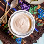 Light orange thick dip is in a small bowl with roasted green peppers on the side. Placed on a colorful brown, blue, green woven tablecloth.