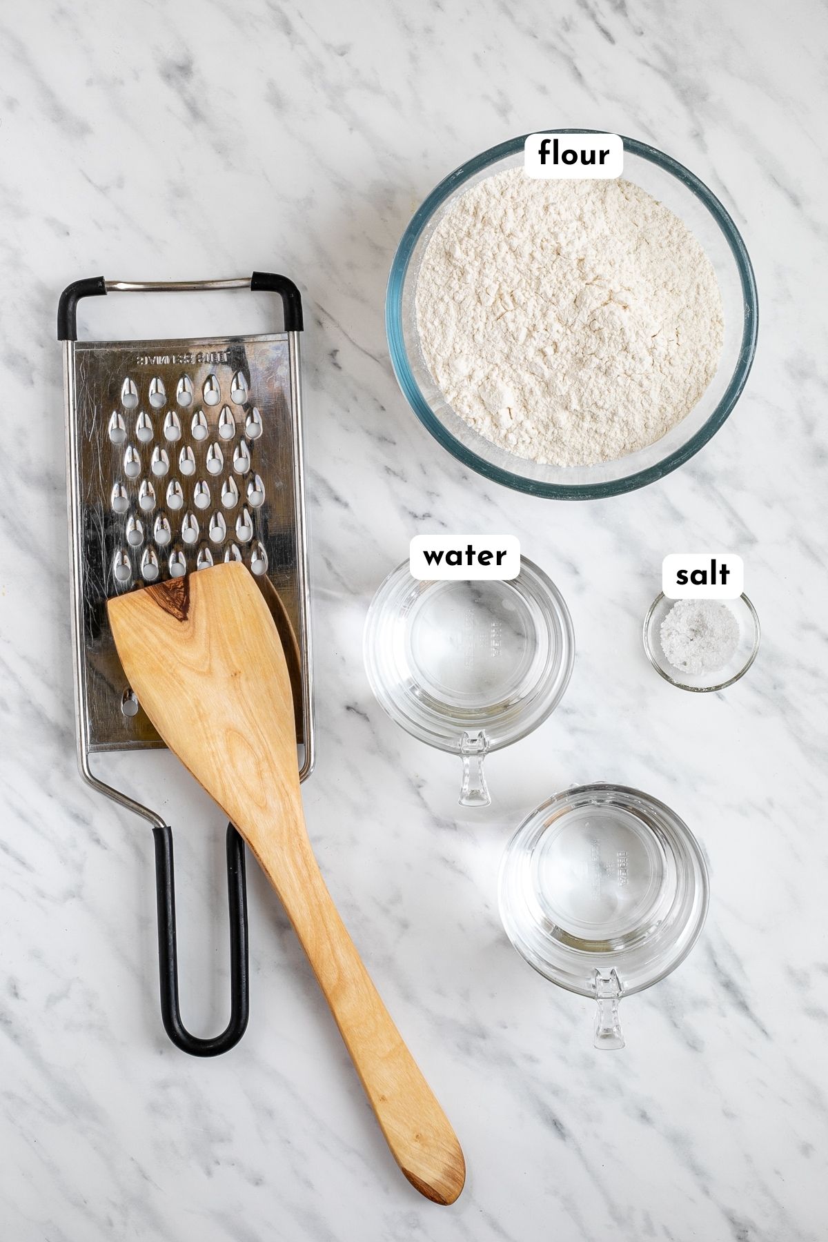 Ingredients of Hungarian dumplings, glass bowl of flour, water and salt with a large hole cheese grater and a wooden spatula at the side.