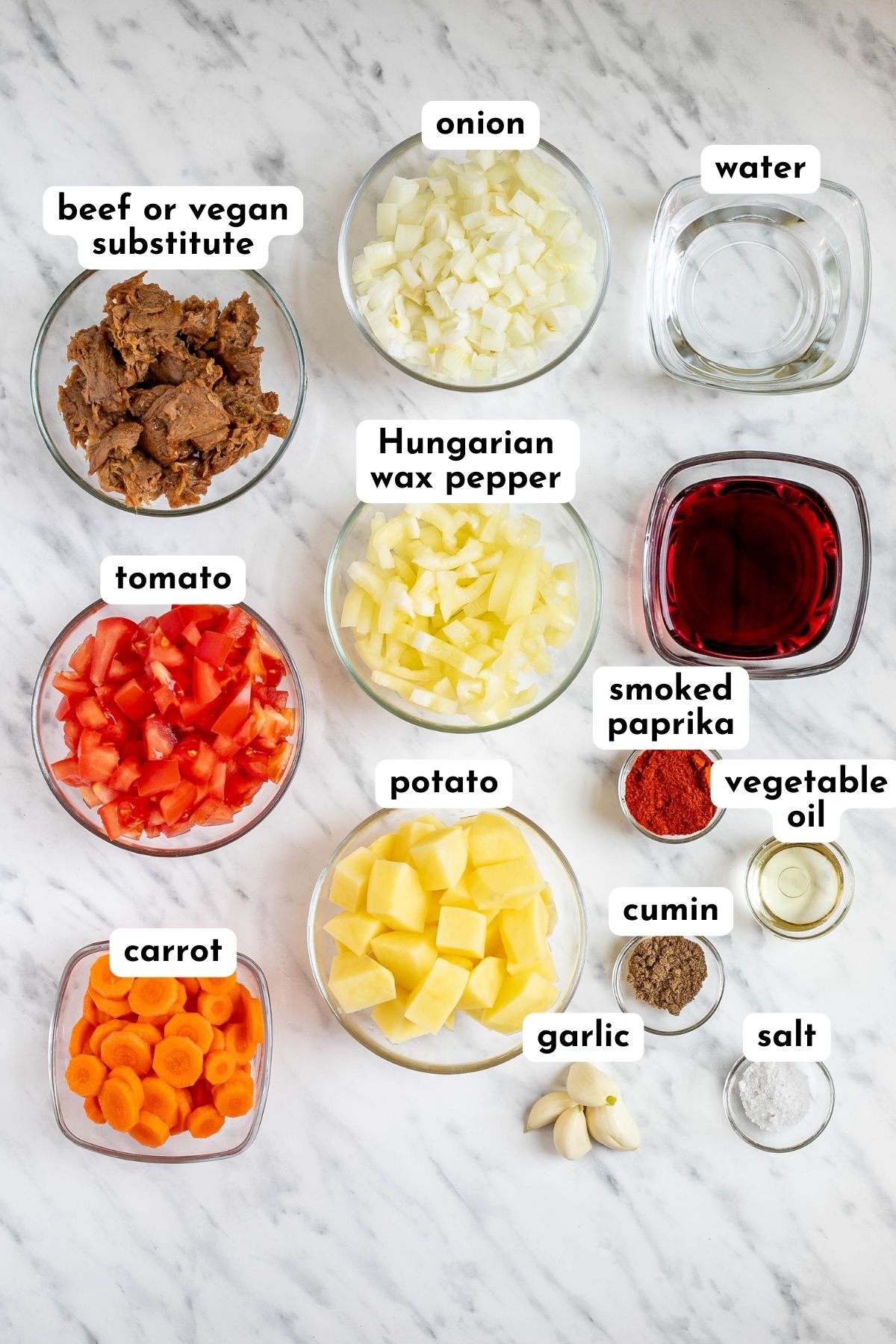Ingredients of Hungarian goulash in small glass bowls like meaty chunks, chopped pepper, tomato, onion, carrot, potatoes, spices, and liquid ingredients.