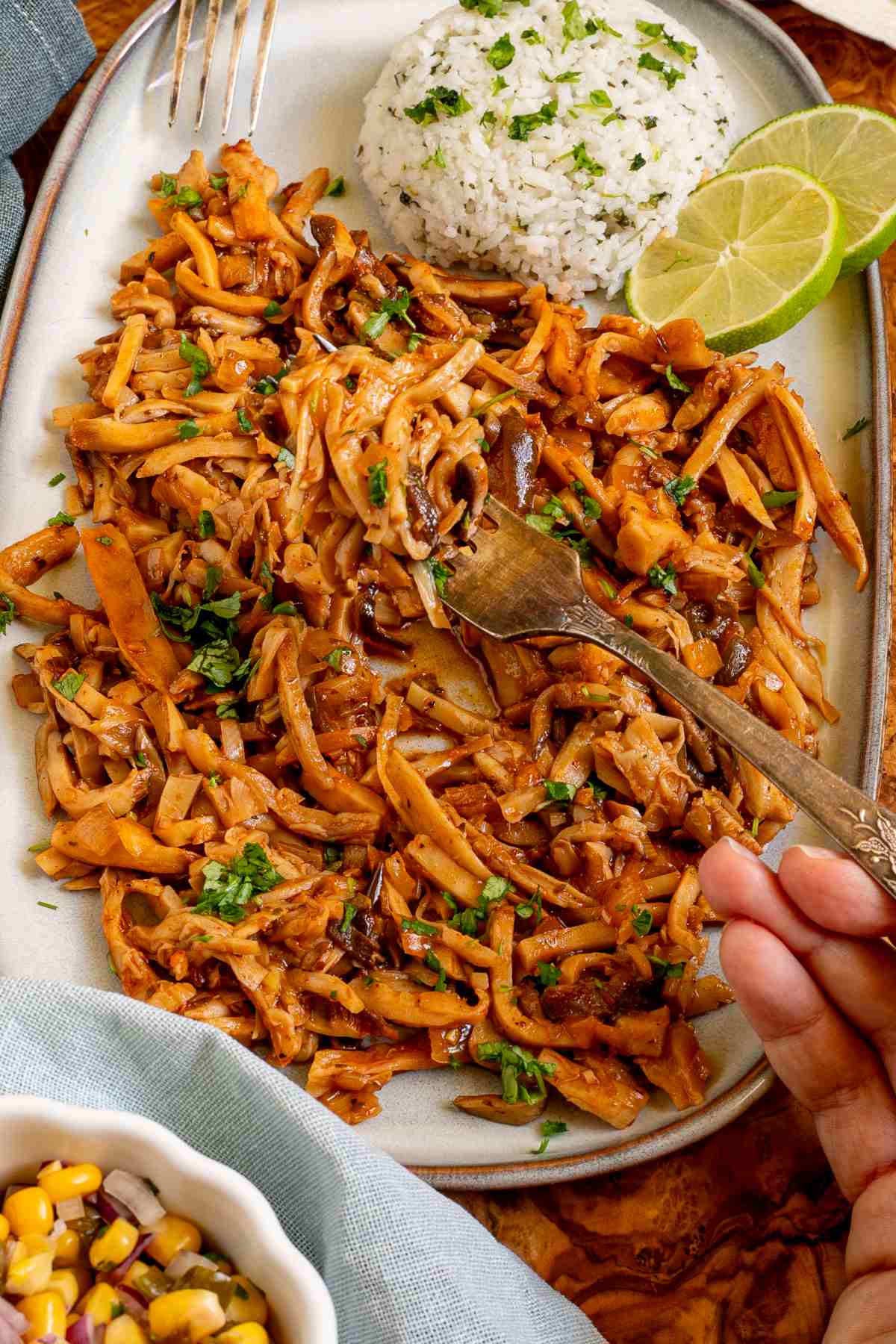 Thin juicy golden brown oyster mushrooms shred sprinkled with chopped green herbs on a light blue tray served with a scoop of rice. A hand is holding a fork to take some.