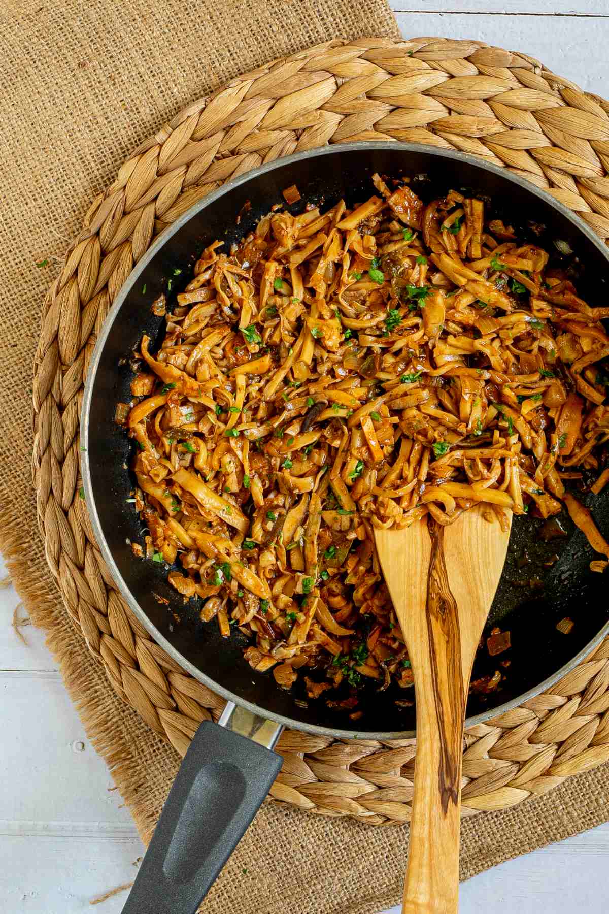 Black frying pan with thin juicy golden brown oyster mushrooms shred sprinkled with chopped green herbs. A wooden spatula is placed inside.