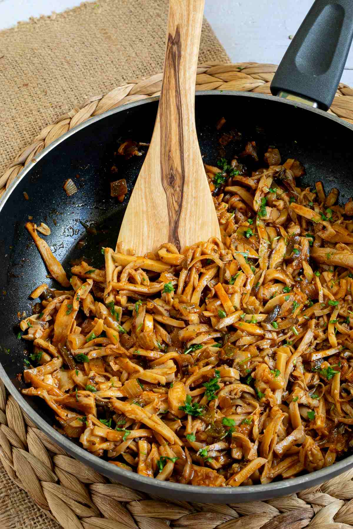 Black frying pan with thin juicy golden brown oyster mushrooms shred sprinkled with chopped green herbs. A wooden spatula is placed inside.