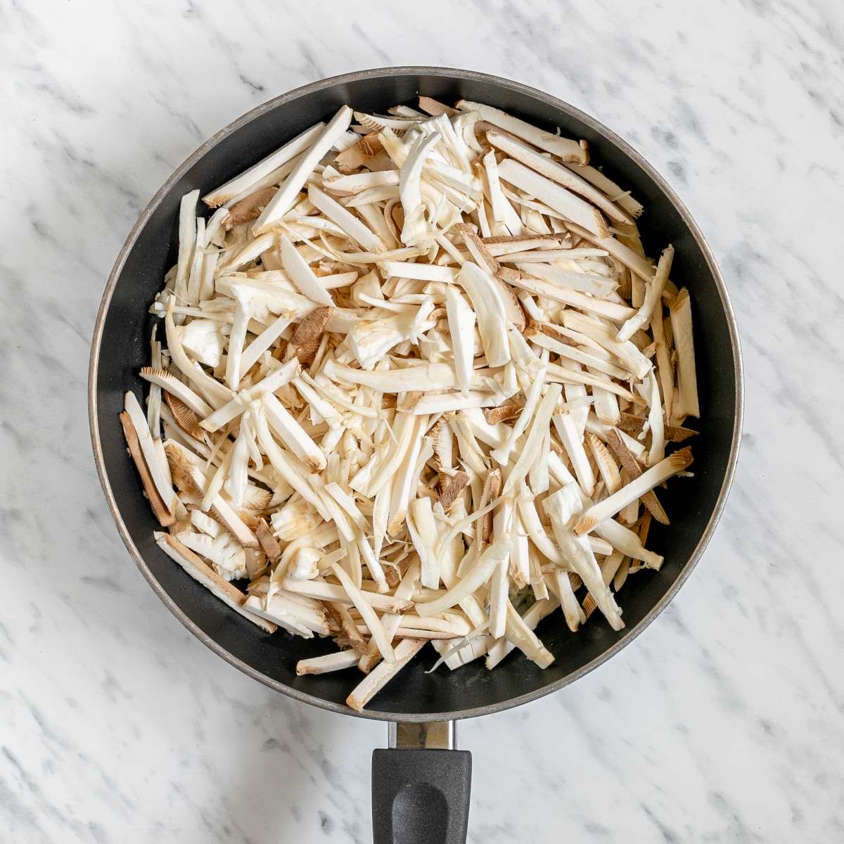 Black frying pan full of thin oyster mushroom slices.