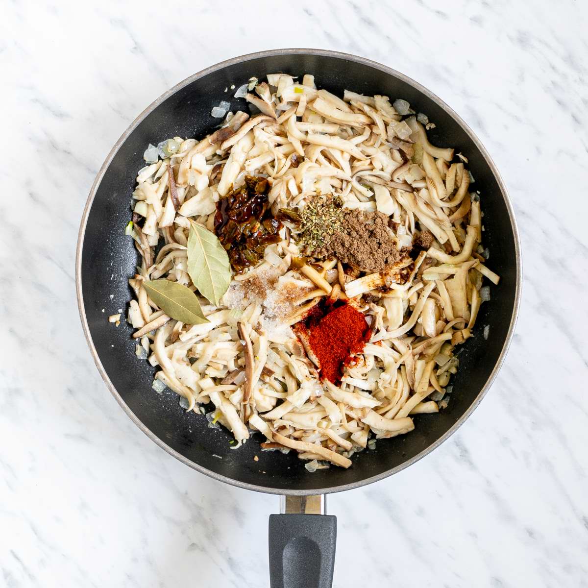 Black frying pan full of thin oyster mushroom slices with small heaps of different spices.