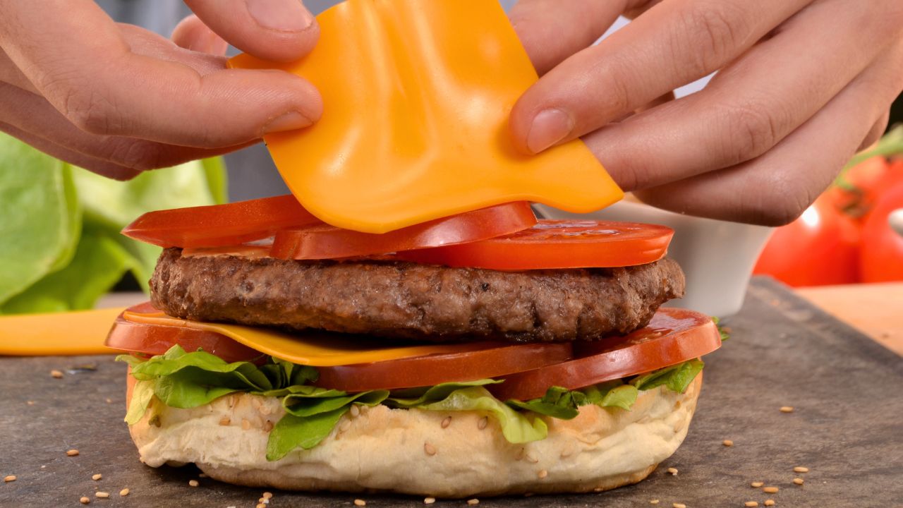 Hands placing a slice of cheese on a layered beef burger with tomatoes and lettuce.