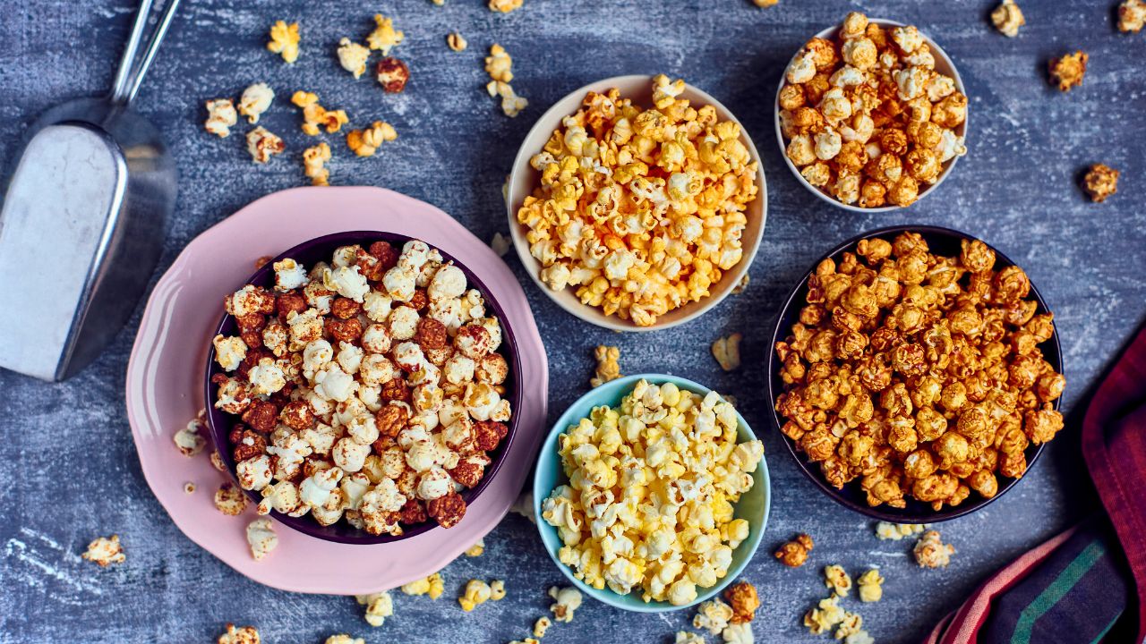 Five bowls of assorted popcorn on a gray surface with scattered kernels.