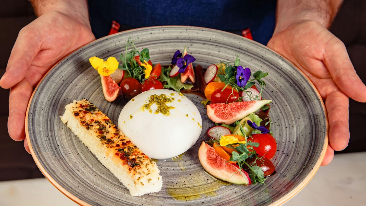 A chef holds a plate with burrata, figs, tomatoes, flowers, and toasted bread.