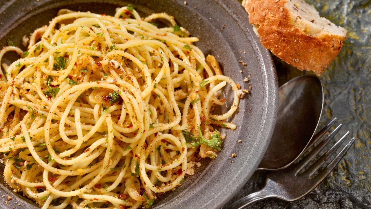 A bowl of spaghetti with herbs and a piece of bread on a dark tabletop.